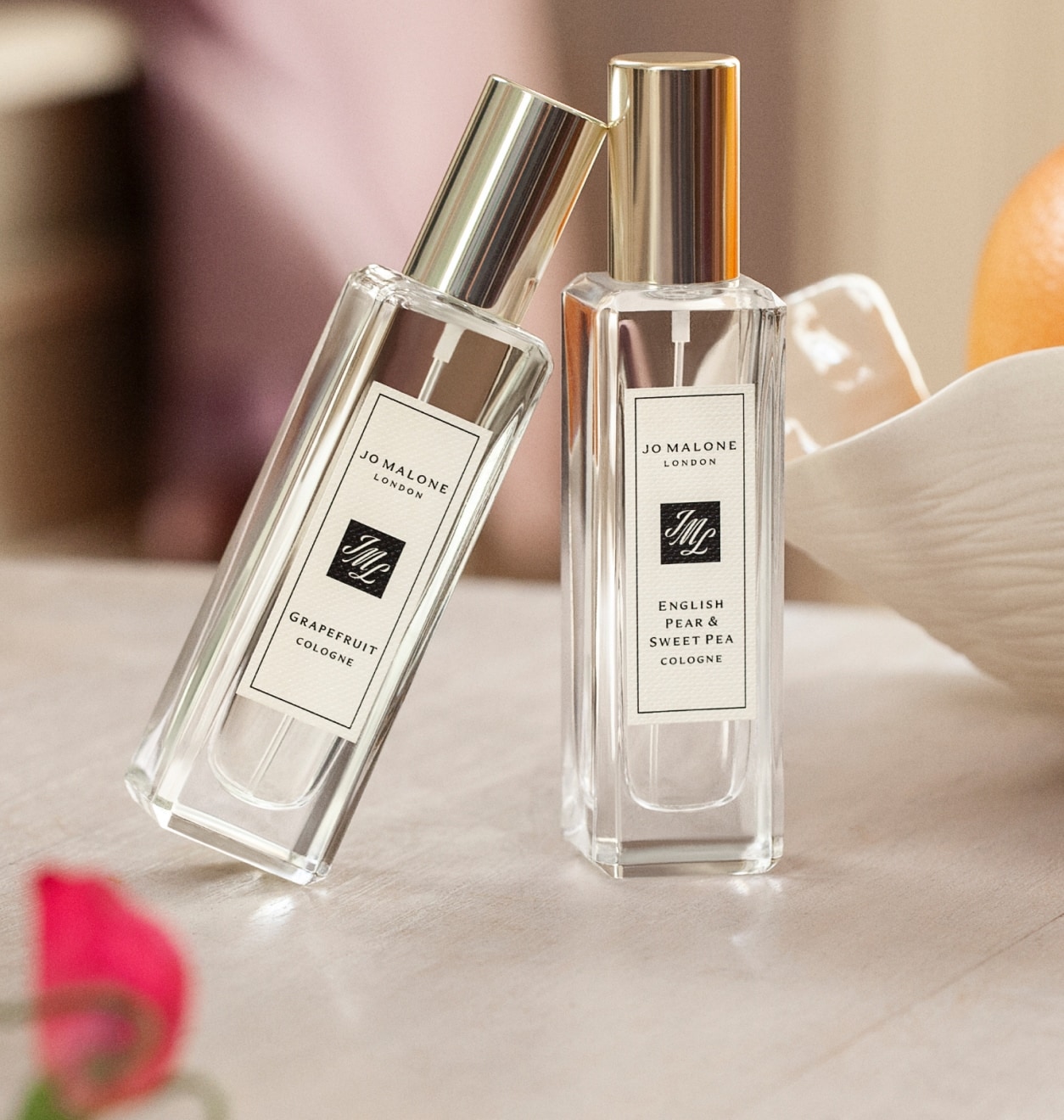 Dos frascos de perfume Jo Malone London con tapones dorados sobre una mesa, junto a un bol de naranjas con pequeñas flores rosas