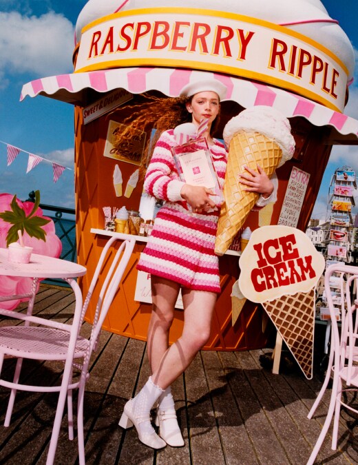 Una persona vestida de rosa sostiene un helado gigante y una fragancia de Jo Malone London en un muelle con asientos rosados.