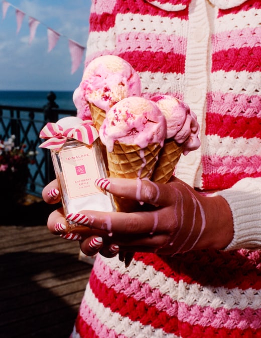Tres conos de helado junto a una fragancia de Jo Malone London con un lazo rosa, en un muelle con vista al mar.