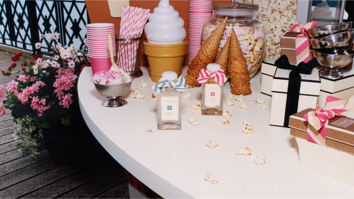 Dos fragancias de Jo Malone London, 1 con lazo azul y otra con lazo rosa, rodeadas de vasos rosados, pajillas, palomitas, dulces