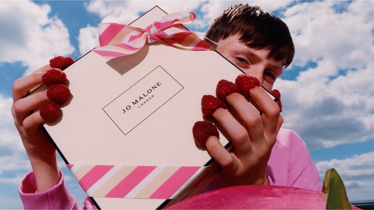 Una caja de Jo Malone London con lazo a rayas rosas es sostenida por una persona que lleva frambuesas en las puntas de los dedos