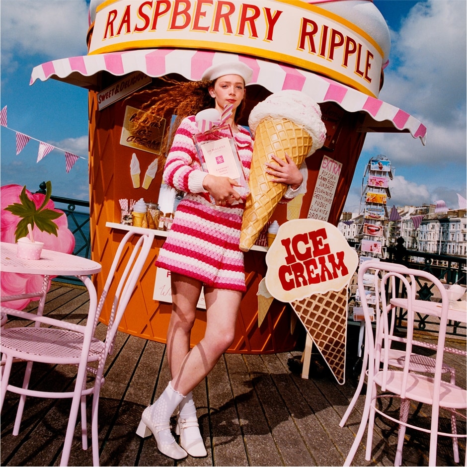 Una persona sostiene un helado gigante y una fragancia de Jo Malone London en un muelle con asientos rosados.