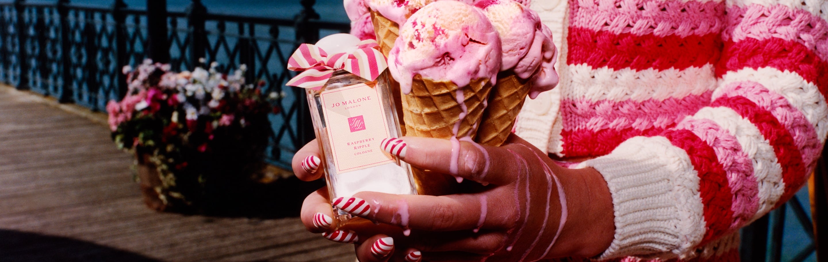 Tres conos de helado junto a una fragancia de Jo Malone London con un lazo rosa, en un muelle con vista al mar.