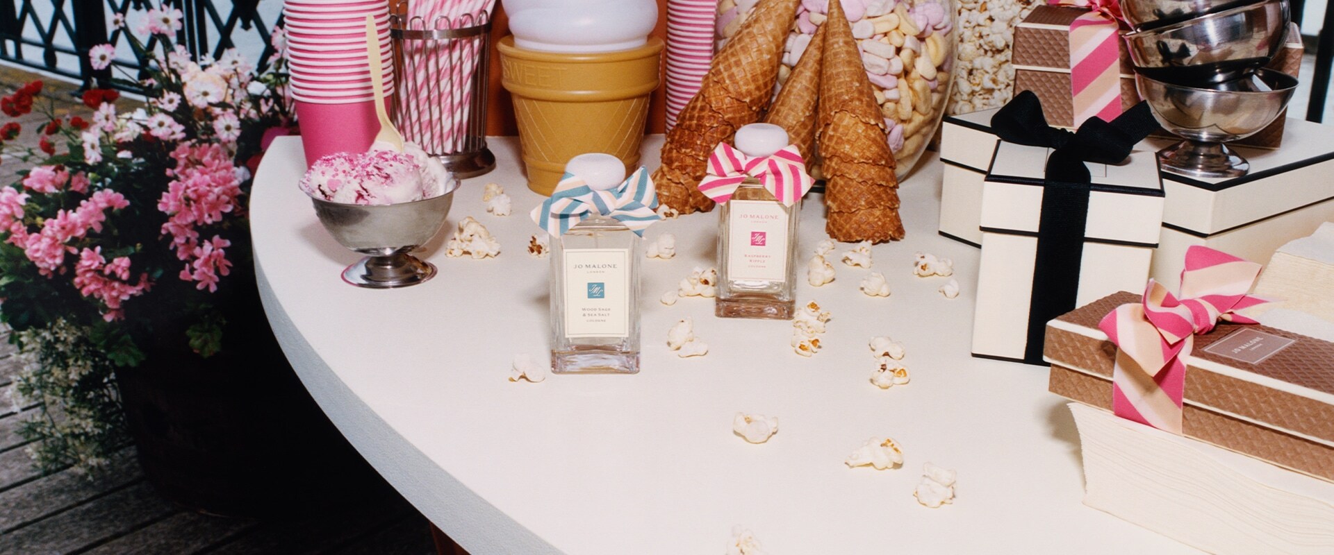 2 fragancias de Jo Malone London, 1 con lazo azul y otra con lazo rosa, rodeadas de vasos rosados, pajillas, palomitas y dulces