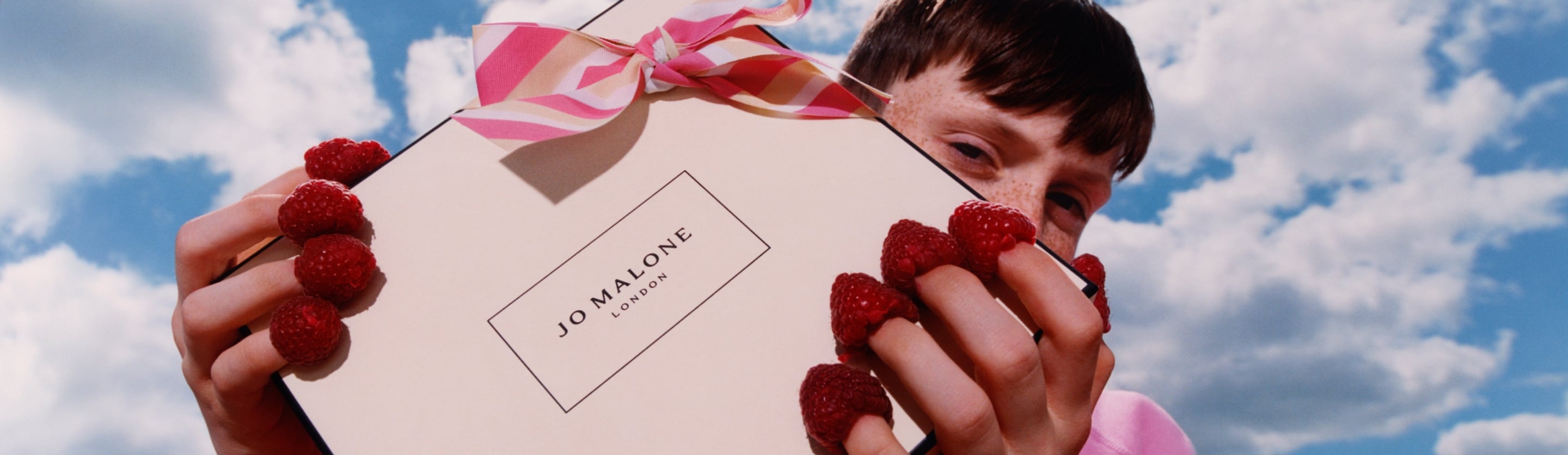Una caja de Jo Malone London con lazo a rayas rosas es sostenida por una persona que lleva frambuesas en las puntas de los dedos