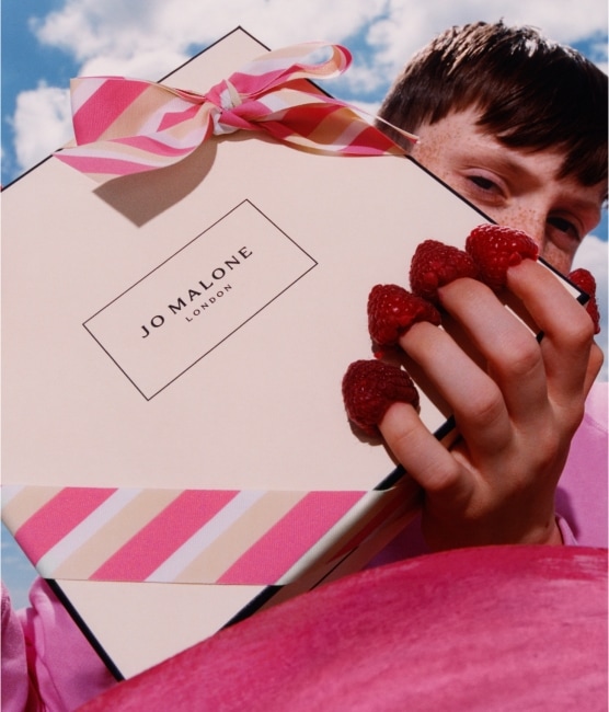 Una caja de Jo Malone London con lazo a rayas rosas es sostenida por una persona que lleva frambuesas en las puntas de los dedos