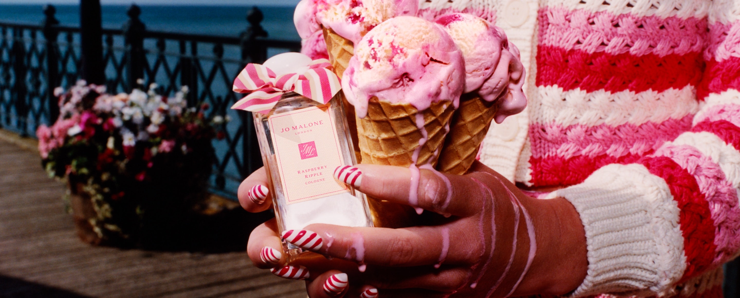 Tres conos de helado junto a una fragancia de Jo Malone London con un lazo rosa, en un muelle con vista al mar.